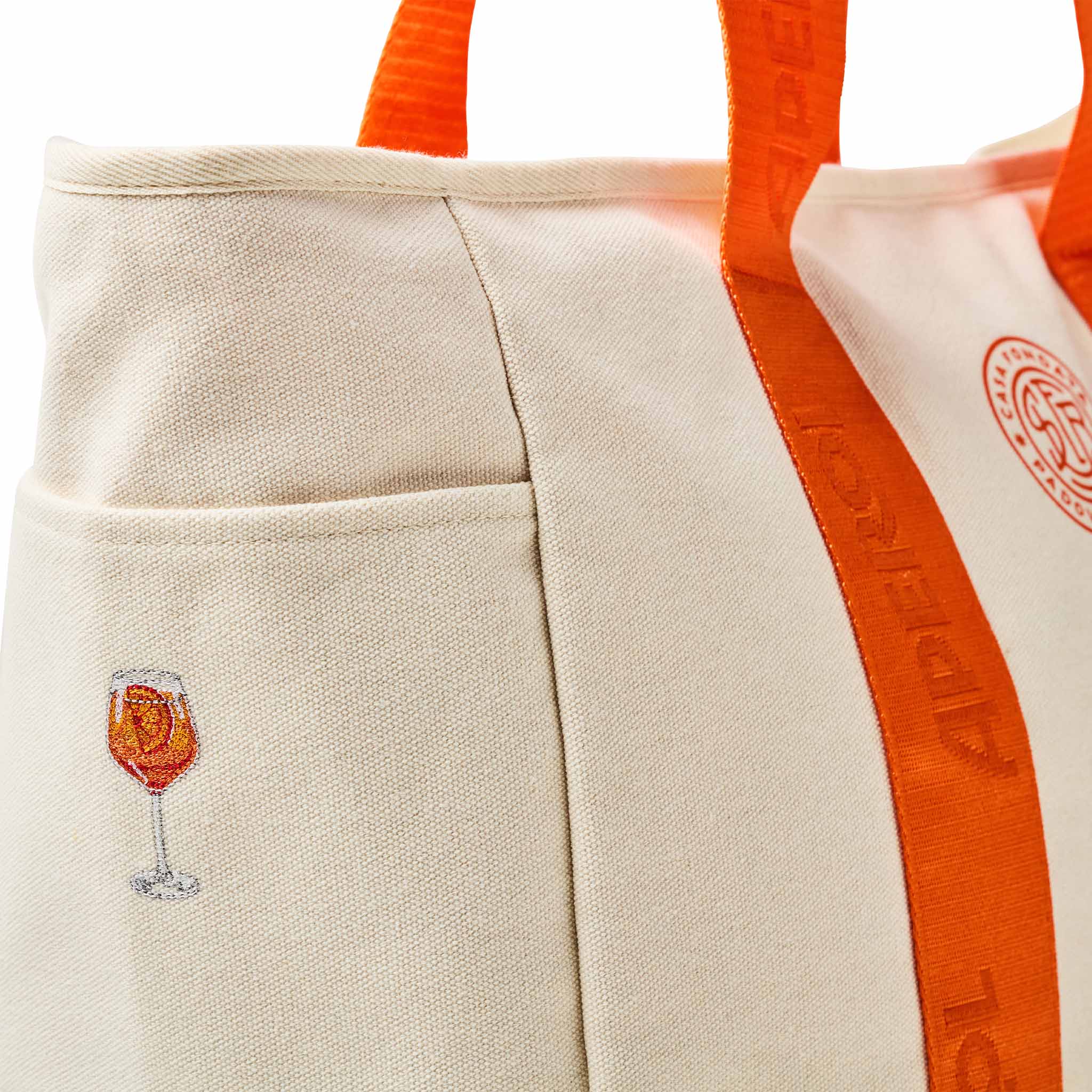 Close-up of a canvas tote bag in warm off-white with bright orange handles and side trim. A small embroidered wine glass with an orange drink appears on the front pocket, and part of a circular orange logo is visible on the side.
