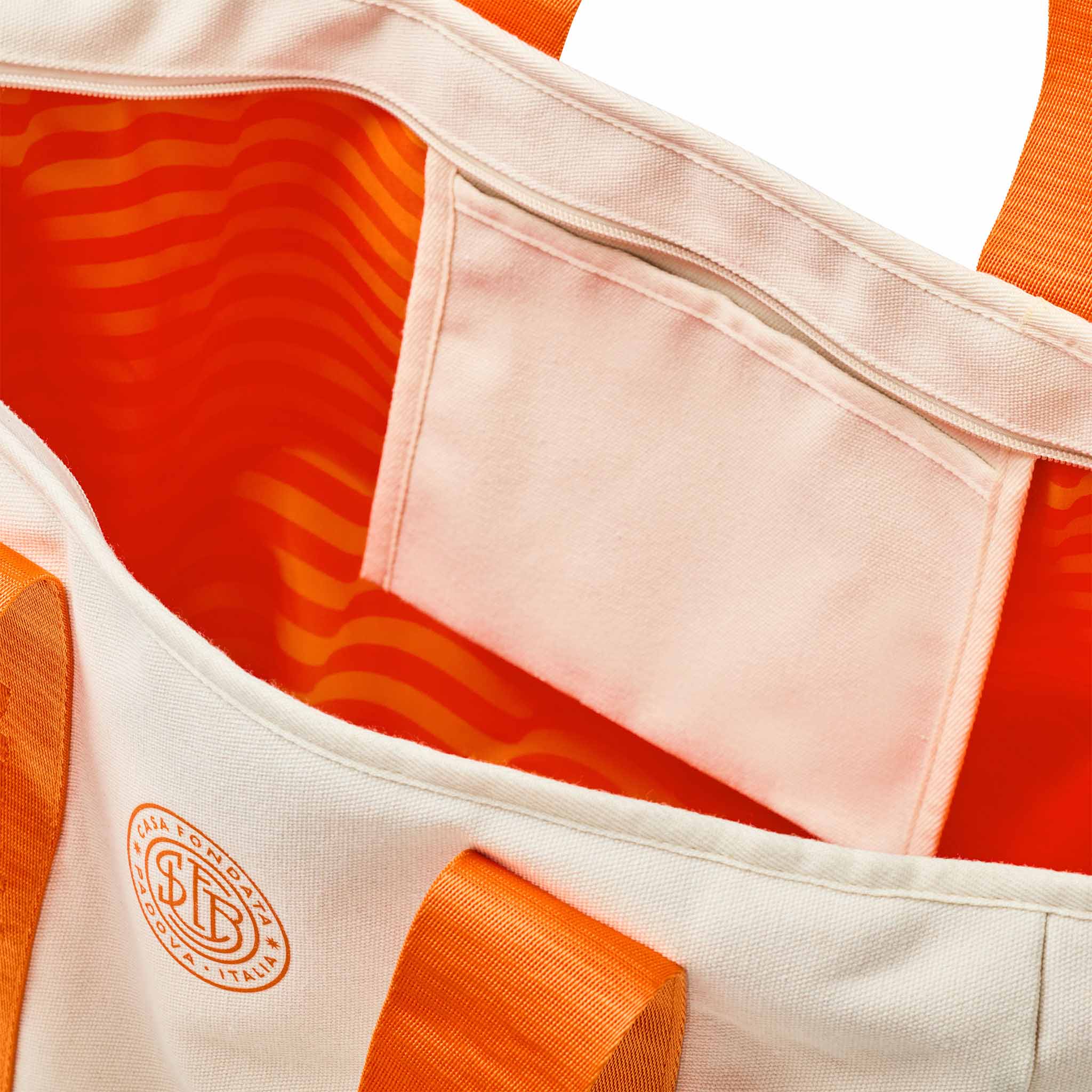 Close-up of a tote bag showing the interior with a peach pocket, bright orange patterned lining, and orange webbing straps. A circular embroidered logo is visible on the front.