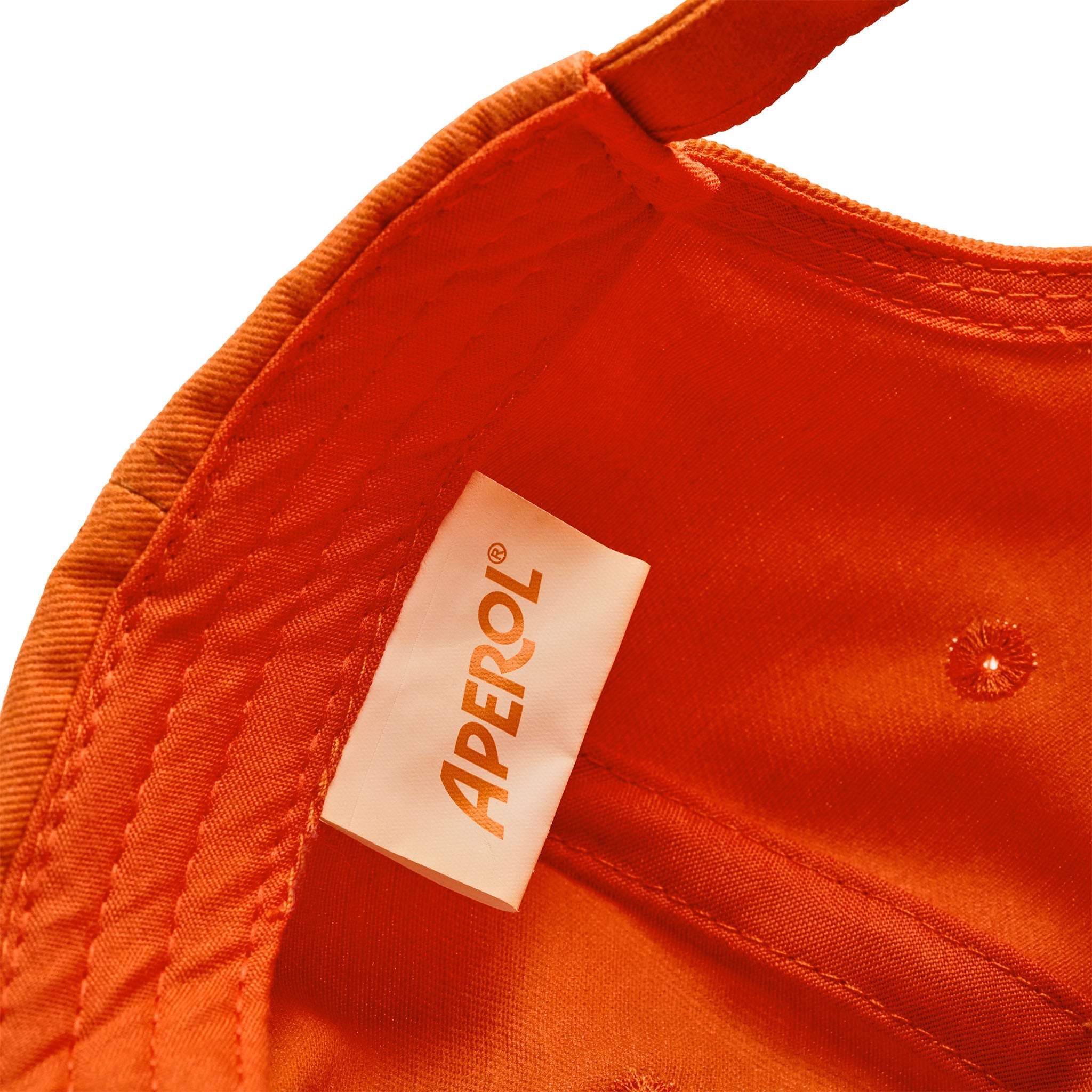 Close-up of an orange fabric product showing a white sewn-in label with the brand name APEROL printed vertically in orange.