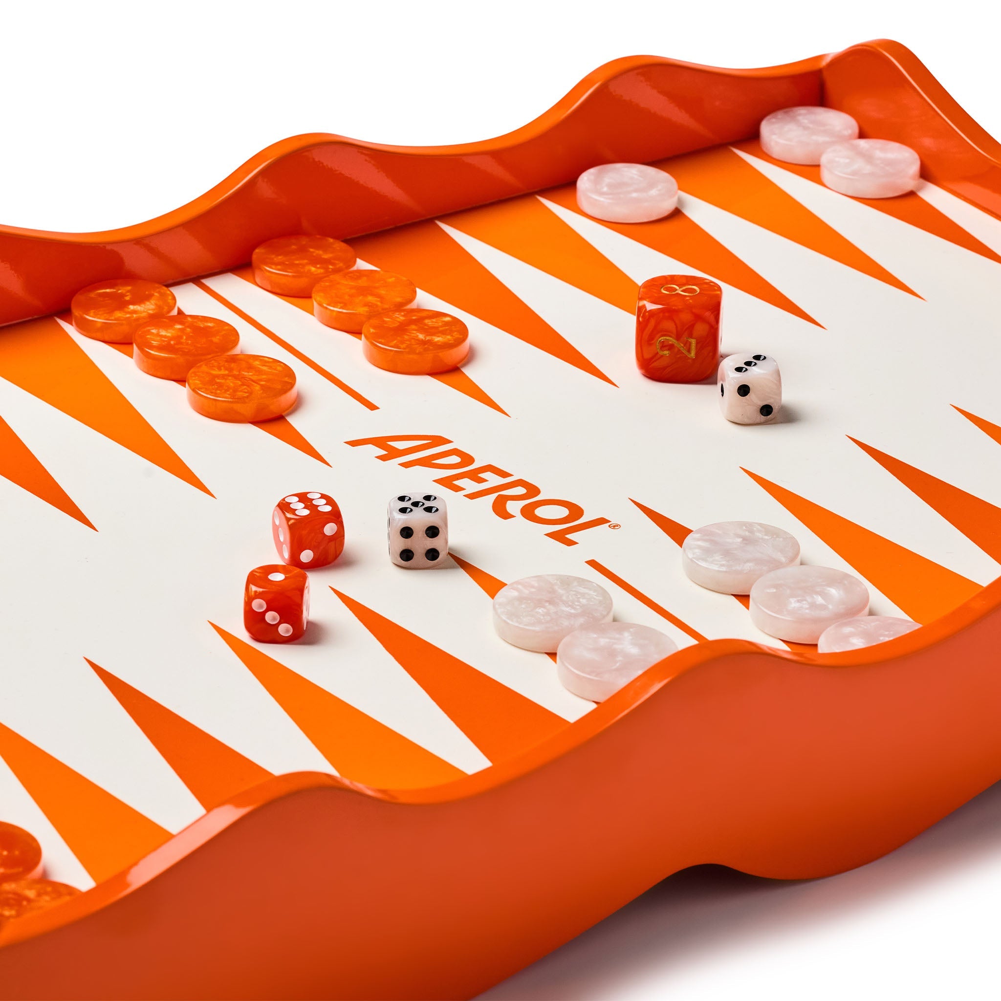 A close-up of a backgammon games tray labeled APEROL, featuring an orange and white triangular track with orange and white playing pieces scattered on it, photographed on a clean white background.