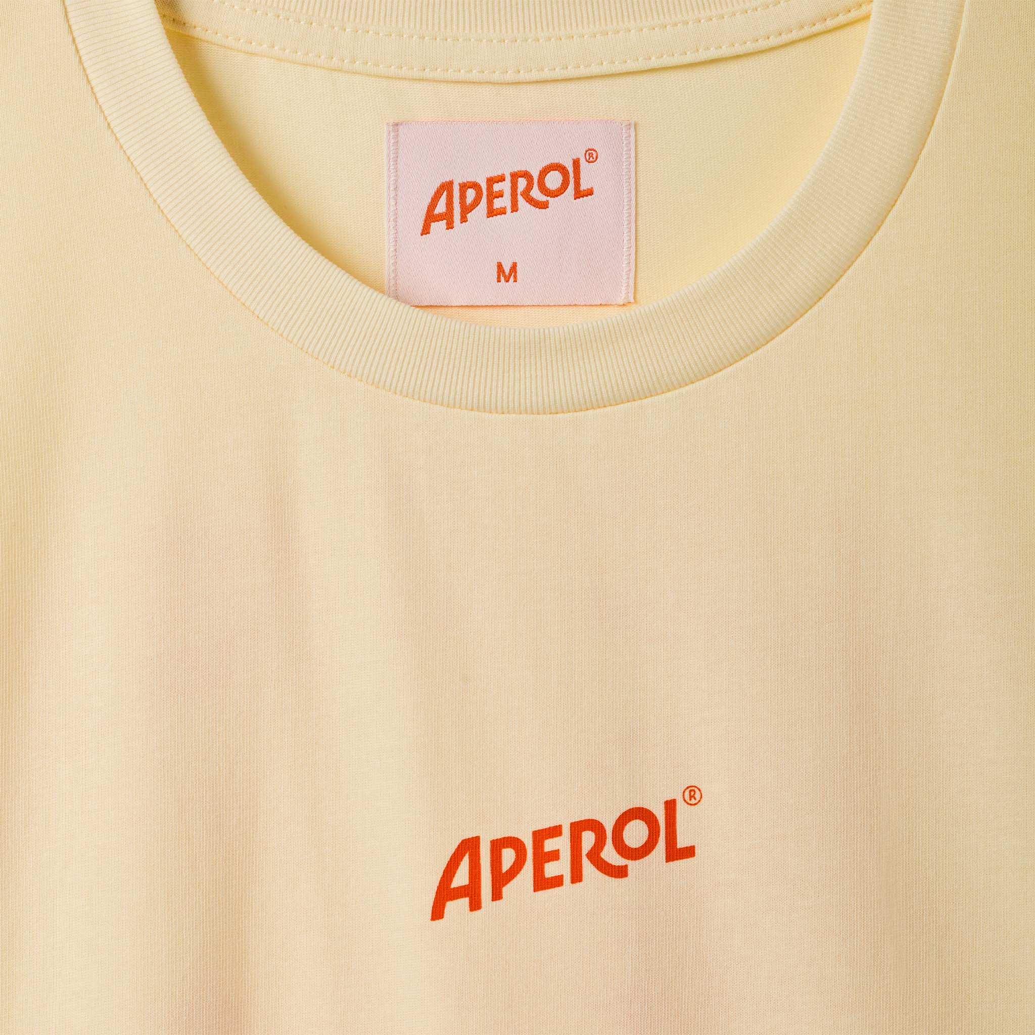 Close-up of a pale yellow crewneck t-shirt with the brand name 'APEROL' printed in orange on the chest and a neck label.