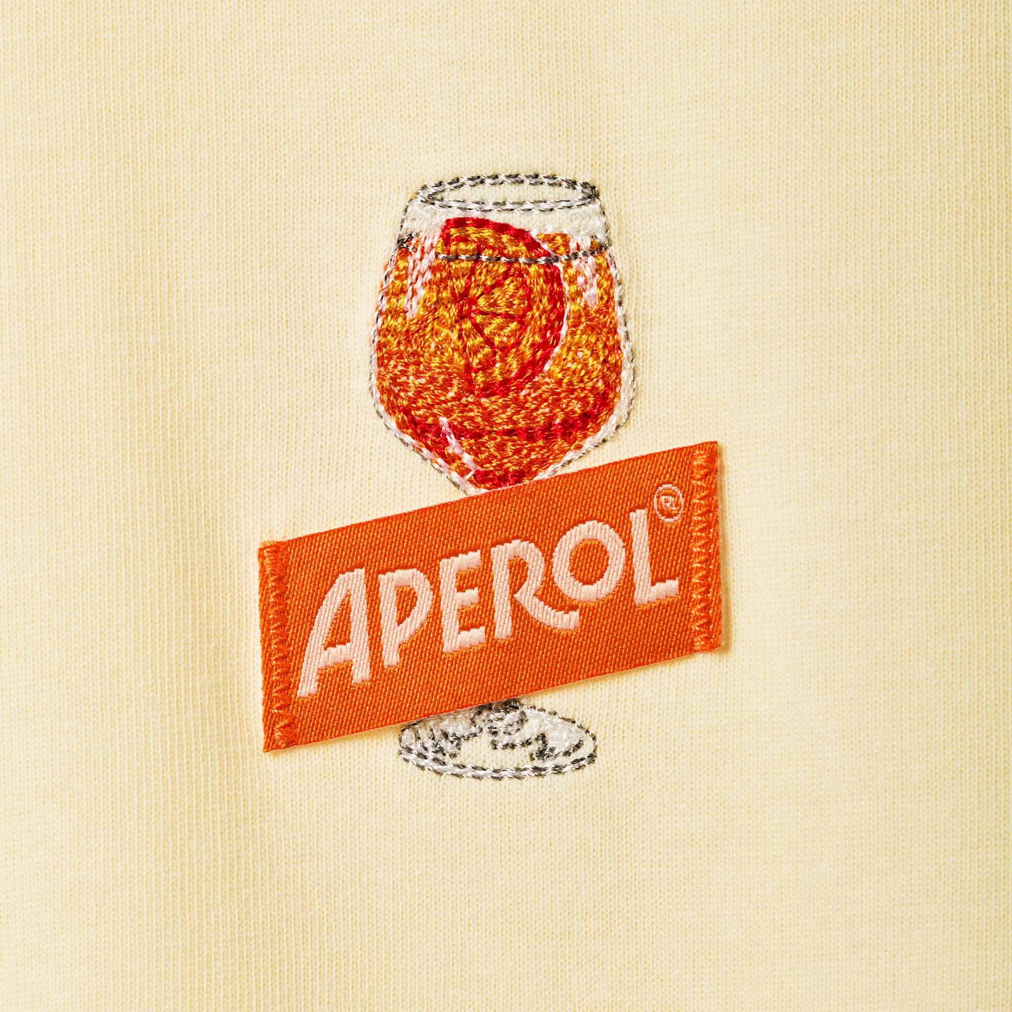 A close-up of a cream-colored woven fabric featuring a prominent embroidered patch of the Aperol brand, depicting an orange cocktail glass with the white 'APEROL' logo on an orange ribbon banner.