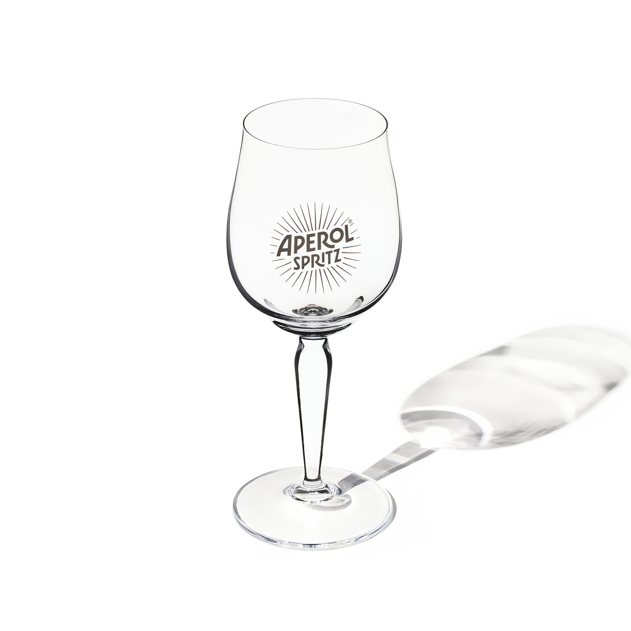A clear aperol spritz glass with the text 'APEROL SPRITZ' and a sunburst design printed on the bowl, shown on a white background with a soft shadow.