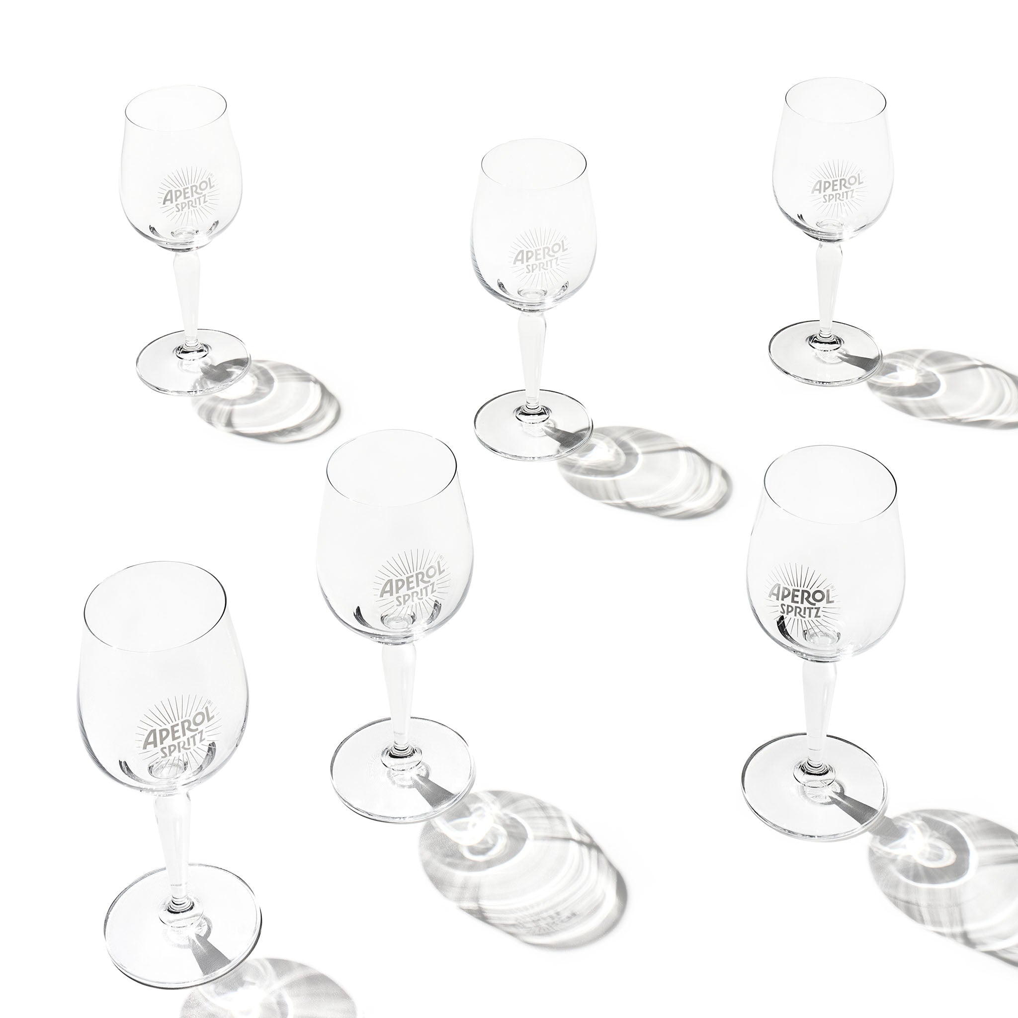 Six clear glasses with 'APEROL SPRITZ' branding, arranged on a white background with soft circular shadows.