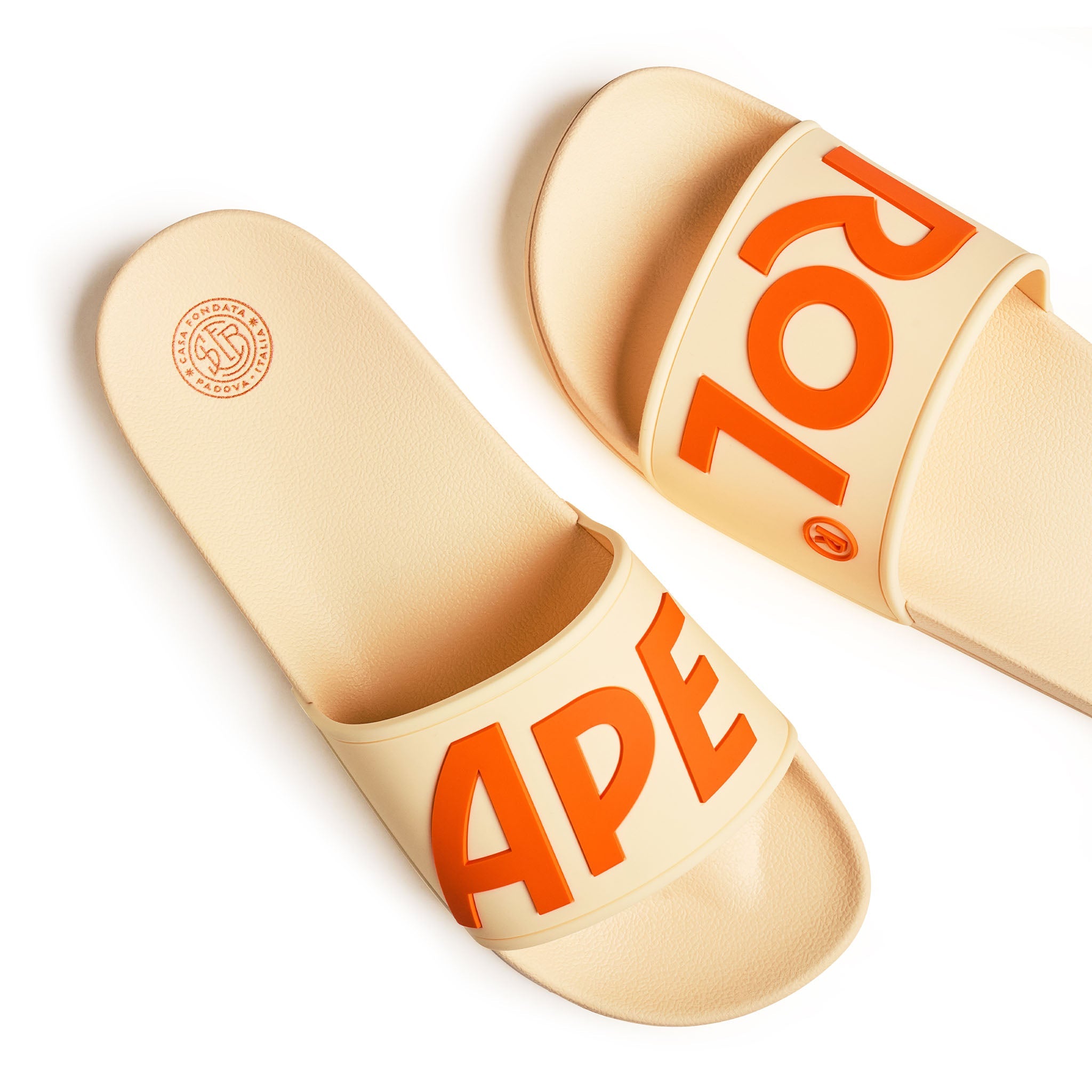 A pair of beige slide sandals with bold orange 'APEROL' lettering on the straps, shown on a clean white background.