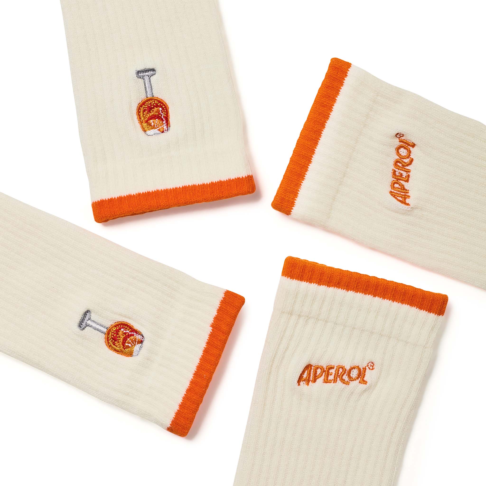 Four cream-colored ribbed socks with bright orange cuffs, arranged on a white background. Two socks feature an embroidered small orange glass graphic, and two feature the word 'APEROL' in orange embroidery.